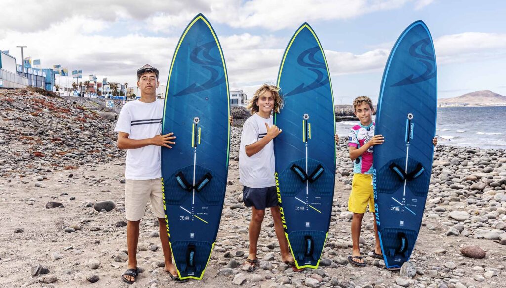 Three young surfers stand on a rocky beach, each holding a blue surfboard with neon-green edges and swimwear/tees.