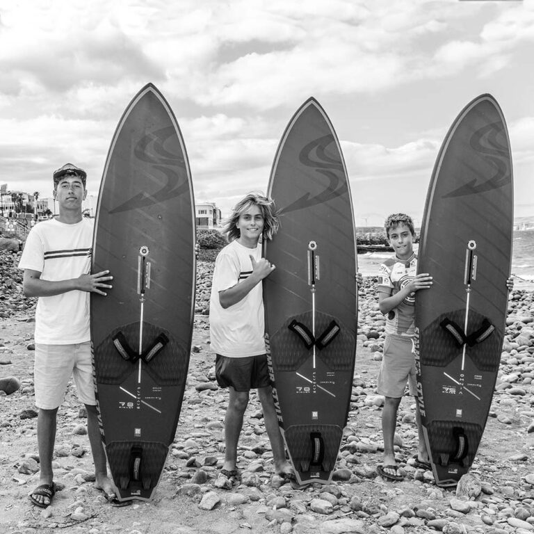 Three teenagers stand on a rocky beach, each holding a large surfboard beside the ocean.