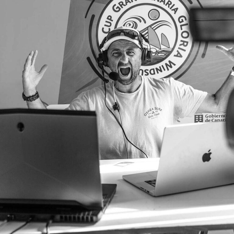 Man wearing a headset and mic shouts into the microphone at a desk with two laptops; a circular event logo is in the background.