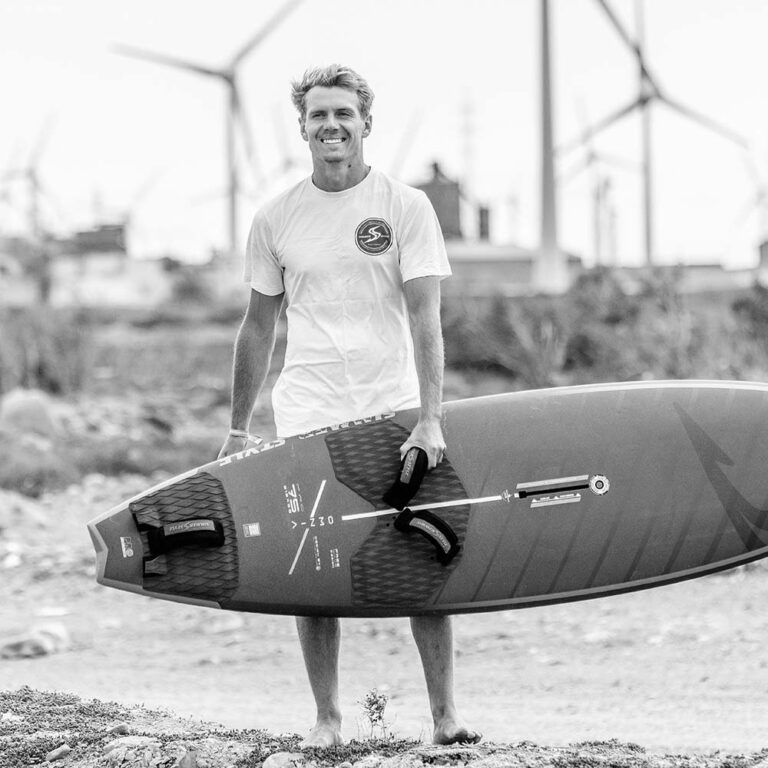 Man standing barefoot on a rocky beach, holding a windsurf board under his arm with harness straps, wind turbines in the background.