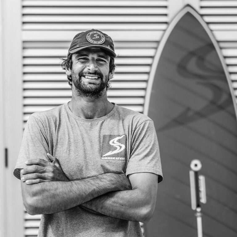 Smiling man in a t-shirt and cap with arms crossed near a surfboard in the background.