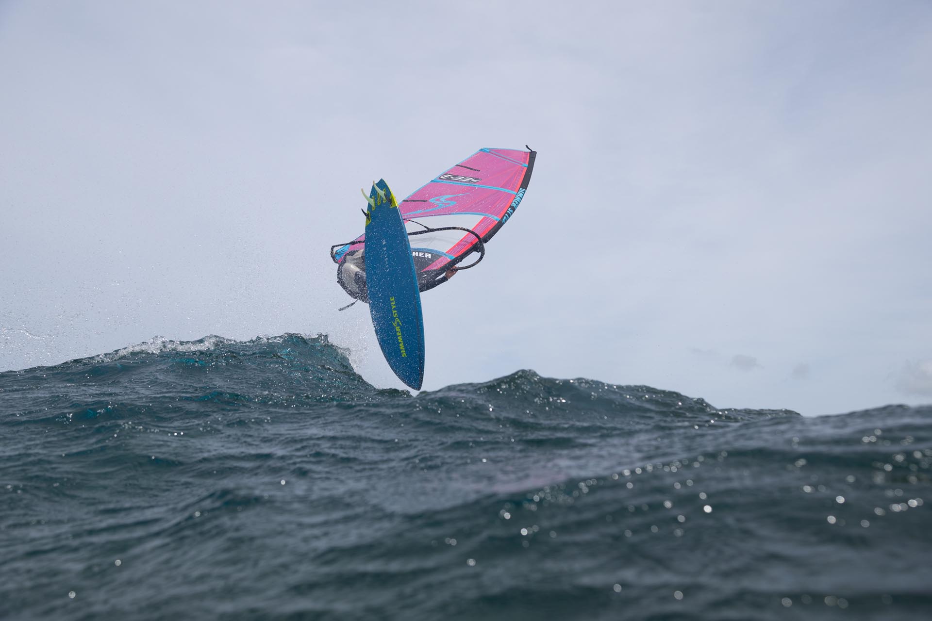 Windsurfer launches off a wave with a pink sail and blue board against a cloudy sky.