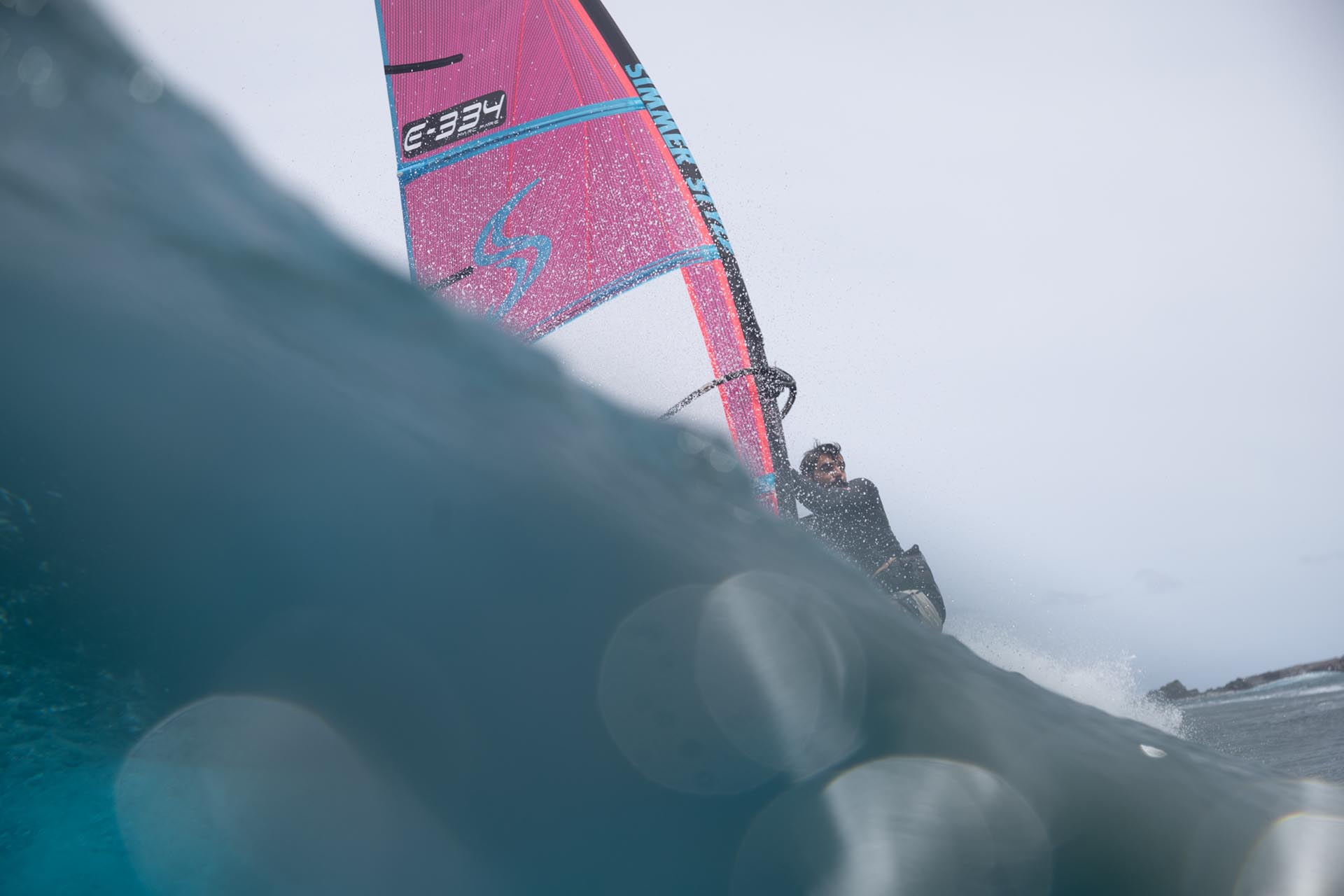 Windsurfer riding a bright pink sail on a cresting wave, viewed from the water with spray around.