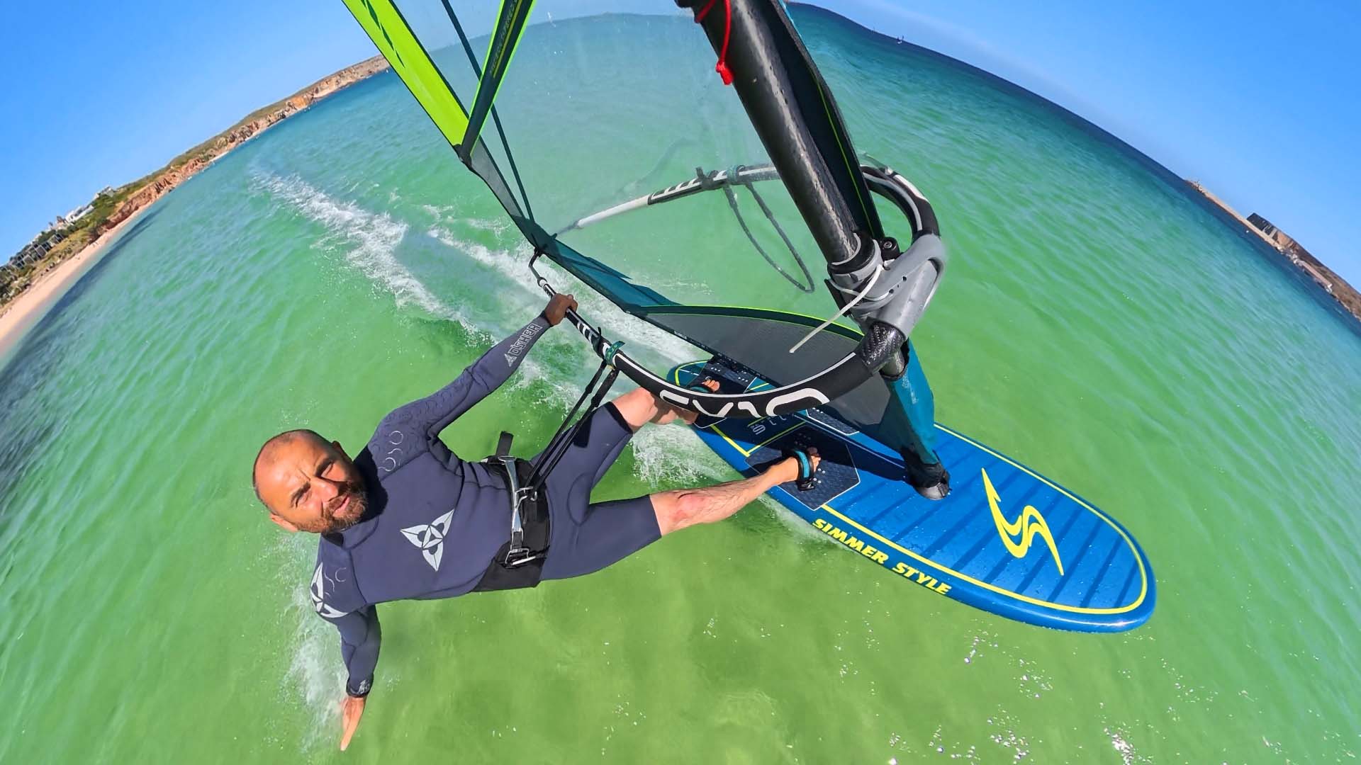 Man windsurfing over turquoise water, grip on the boom, blue and yellow board slicing through the Bay water on a sunny day.