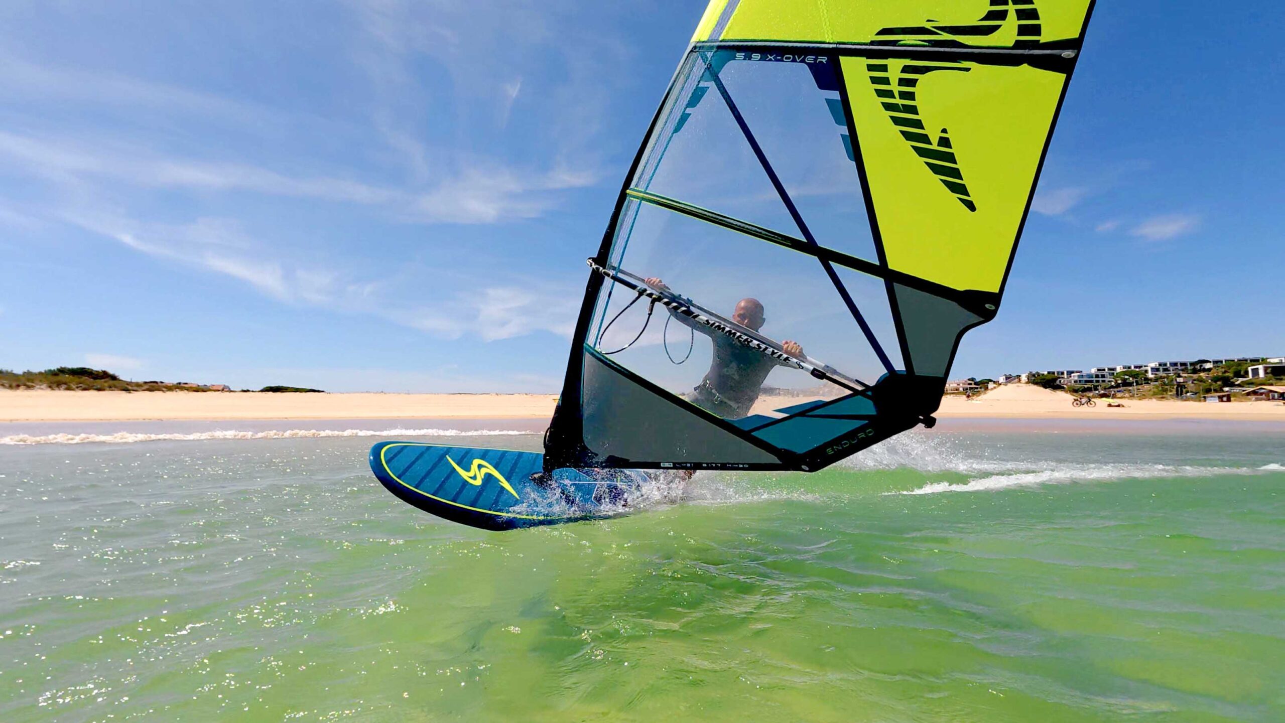 Windsurfer riding a turquoise-green wave with a bright yellow sail and a sandy beach in the background.