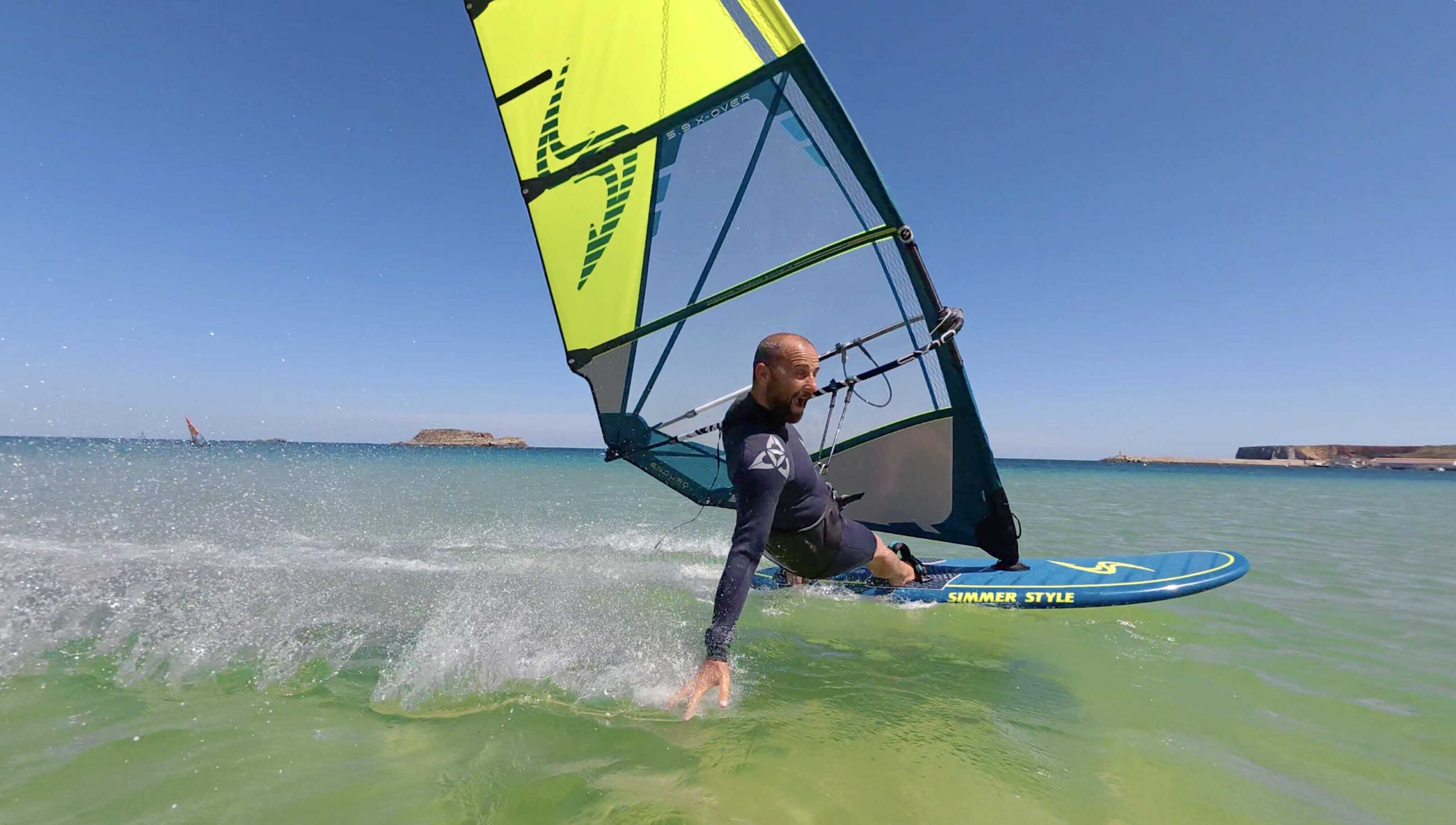 Windsurfer riding a board on turquoise water with a bright yellow sail, spray splashing around.