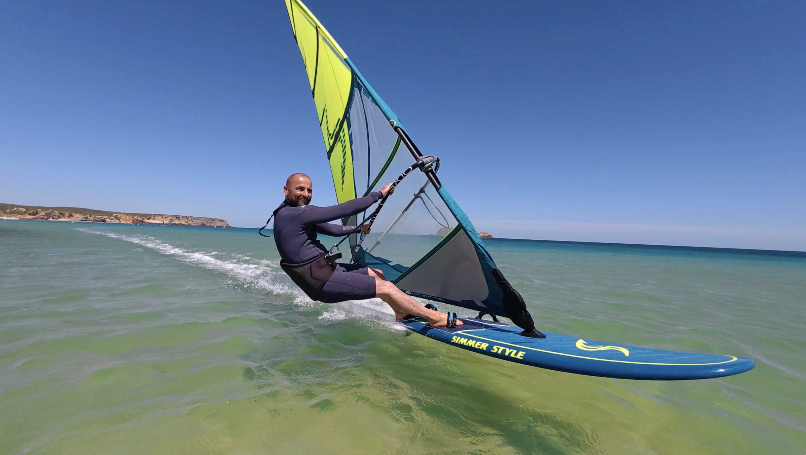 Man windsurfing on calm sea with a bright yellow sail and turquoise water, rocky coast in the distance.