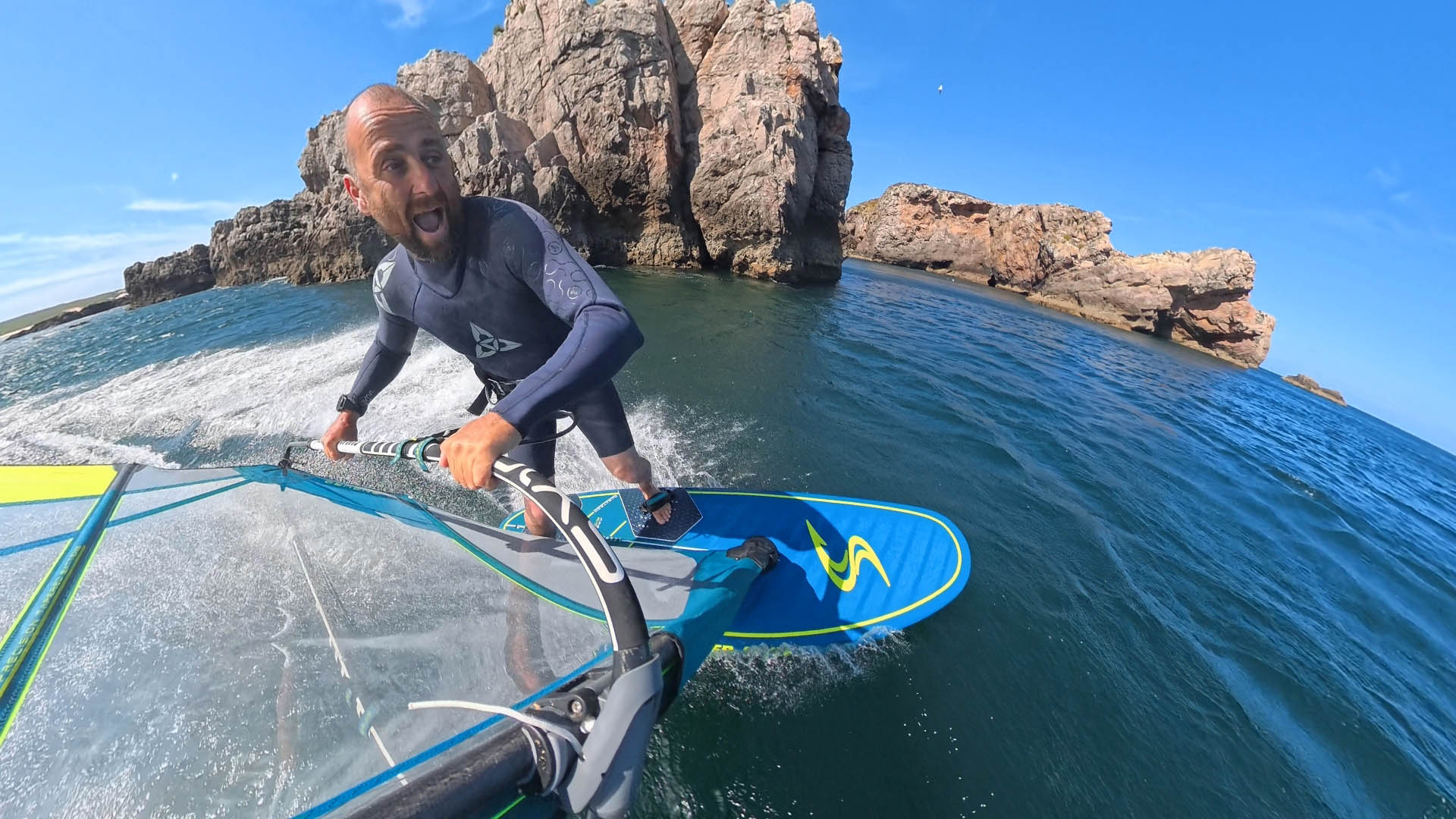 Man windsurfing on a blue board, gripping the sail in a wetsuit with rocky cliffs in the background and a bright blue sky, smiling.
