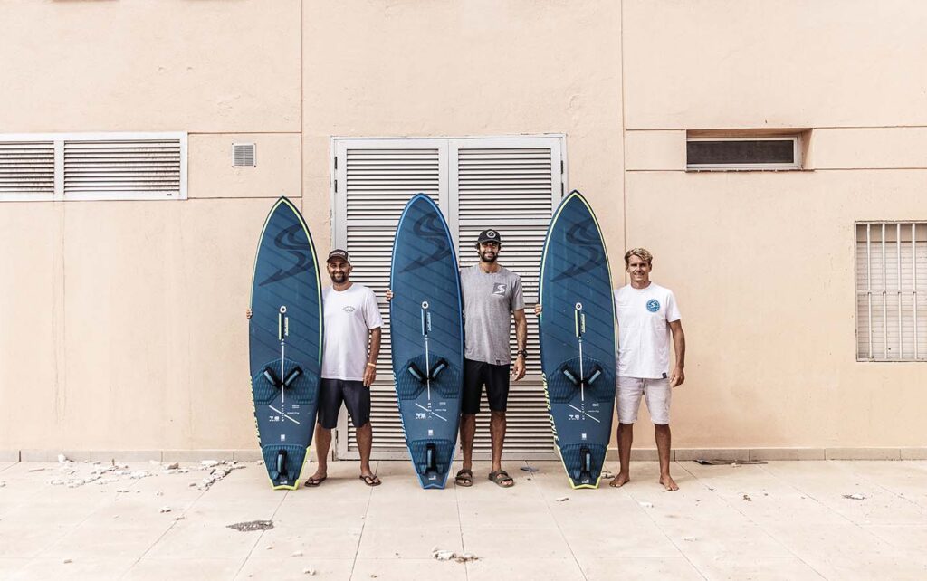 Three men standing in a row, each holding a blue surfboard, in front of a beige building with windows.