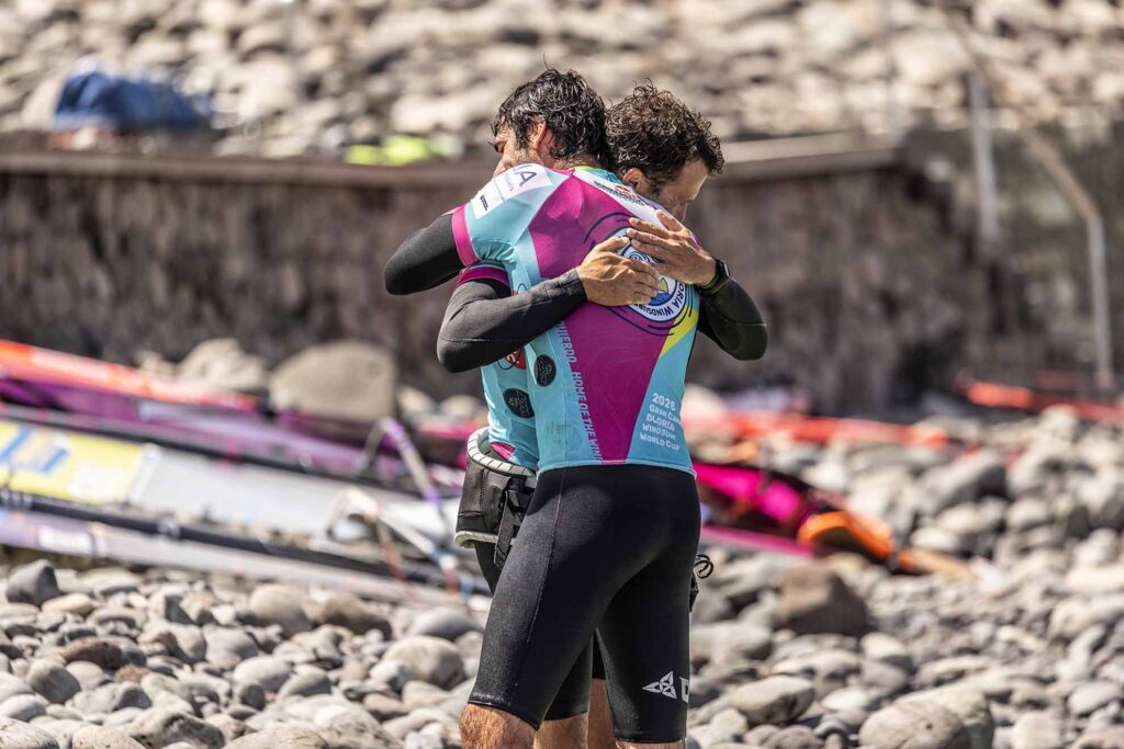 Two athletes share a hug during a sporting event by a rocky shoreline, wearing bright turquoise and pink jerseys written with event logos.