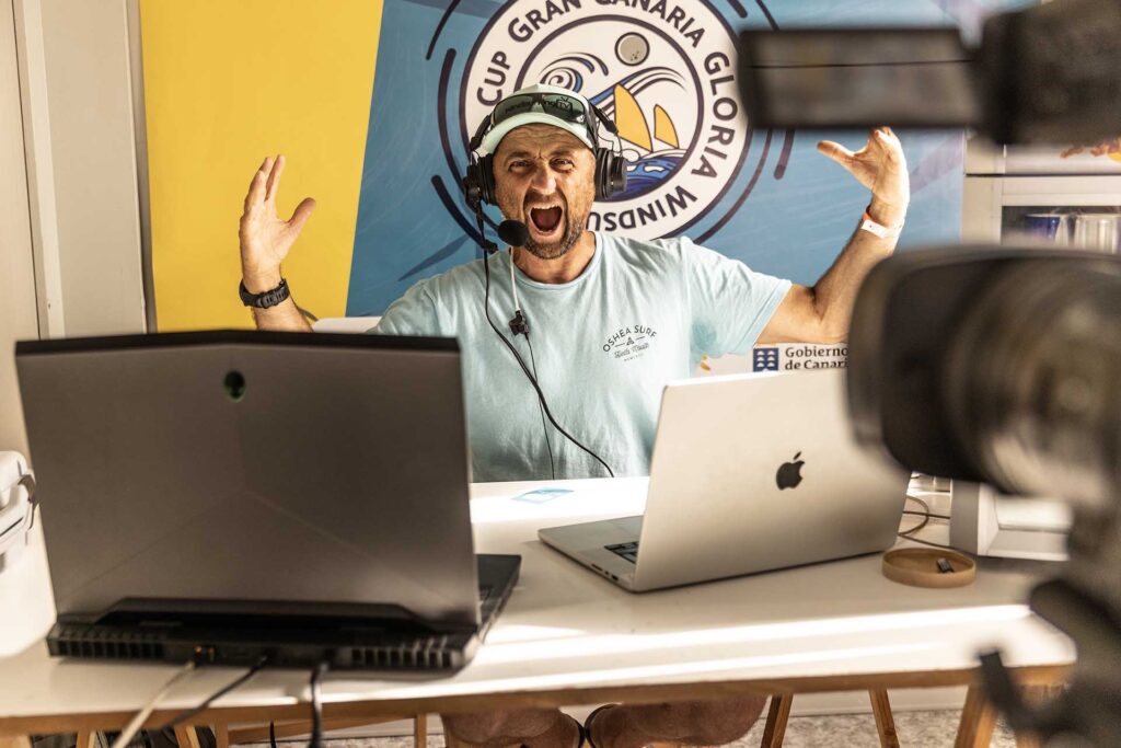 Man wearing a headset and microphone screams into the mic while raising hands at a broadcasting desk, with colorful banner behind him.