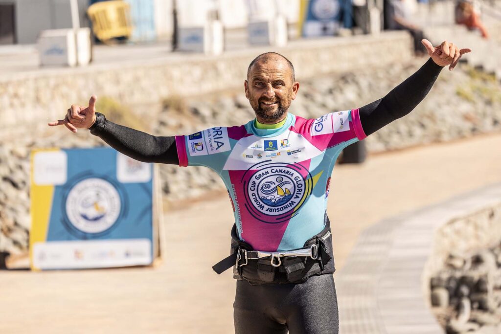 Male cyclist in a colorful race jersey raises both arms in celebration on a seaside promenade.