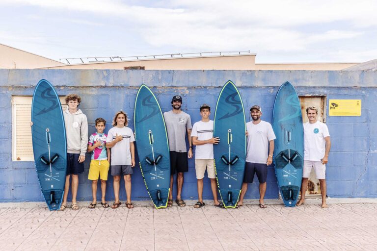 Eight people of varying ages stand side by side, each holding a blue surfboard in front of a weathered blue wall at the beach.