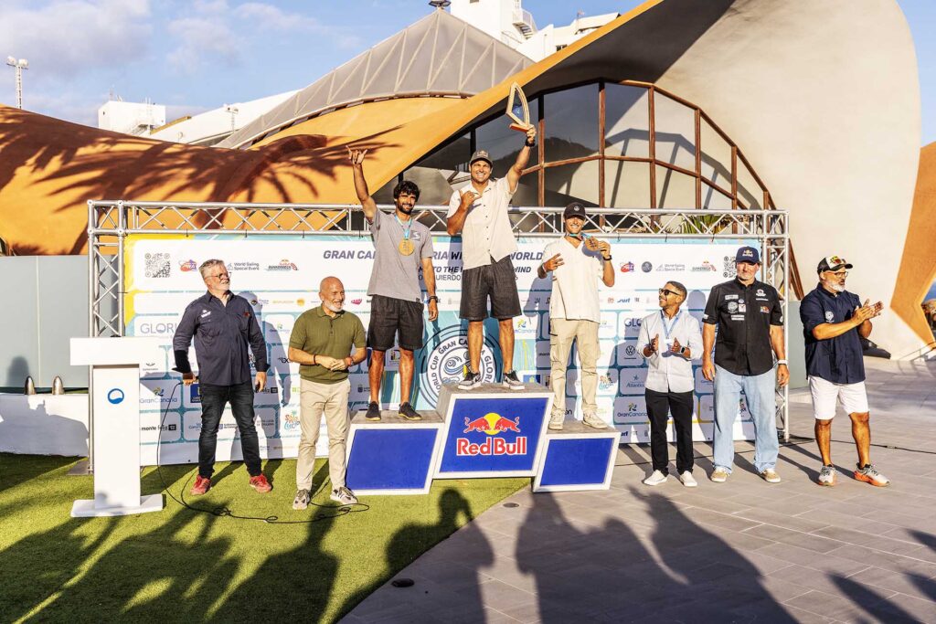 Three men stand on a prize podium posing with medals and a trophy as officials applaud at a sailing event, Red Bull branding visible on the podium guard.