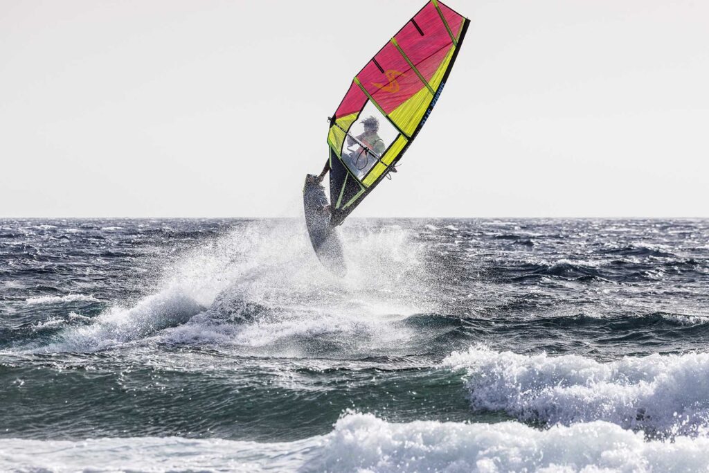 Windsurfer airborne above blue ocean waves with bright pink-yellow sail.