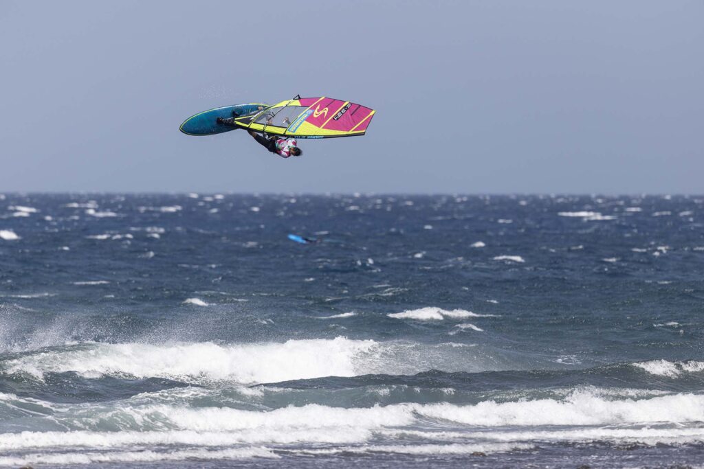 Windsurfer leaps above white-capped waves with a bright pink-yellow sail, sea and blue sky in the background, spray trailing from the board.