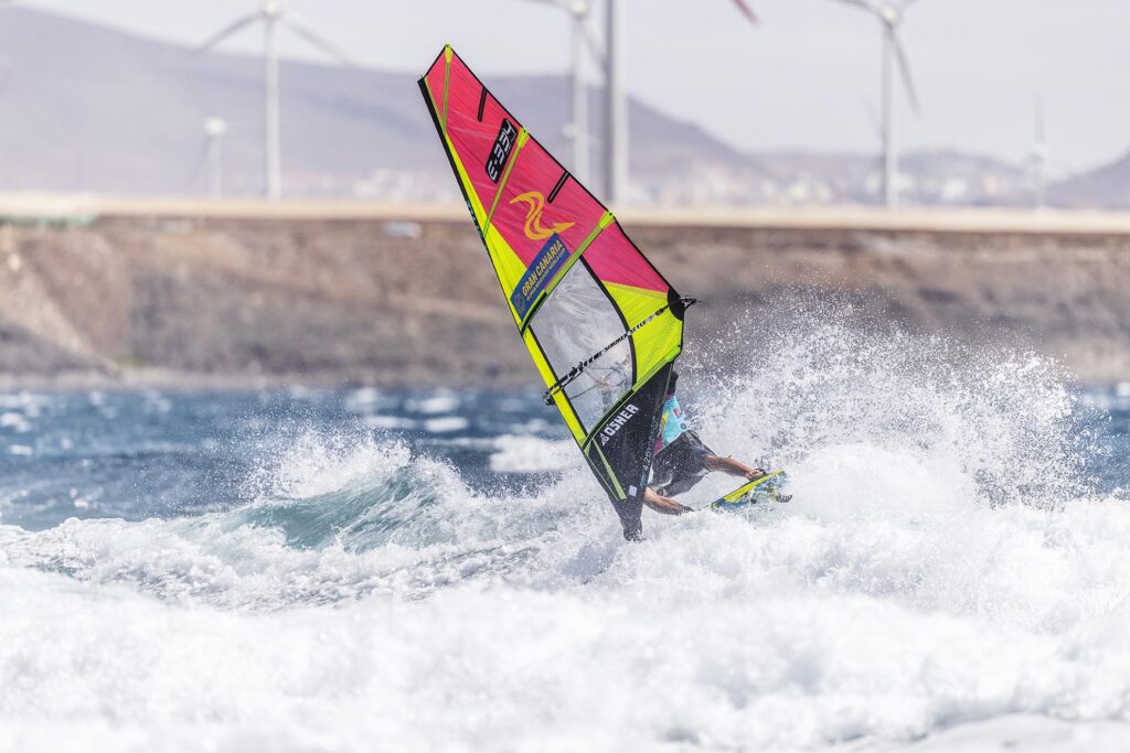 Wind surfer launching off a wave with a bright pink and neon yellow sail as spray erupts, coastal backdrop in the distance.