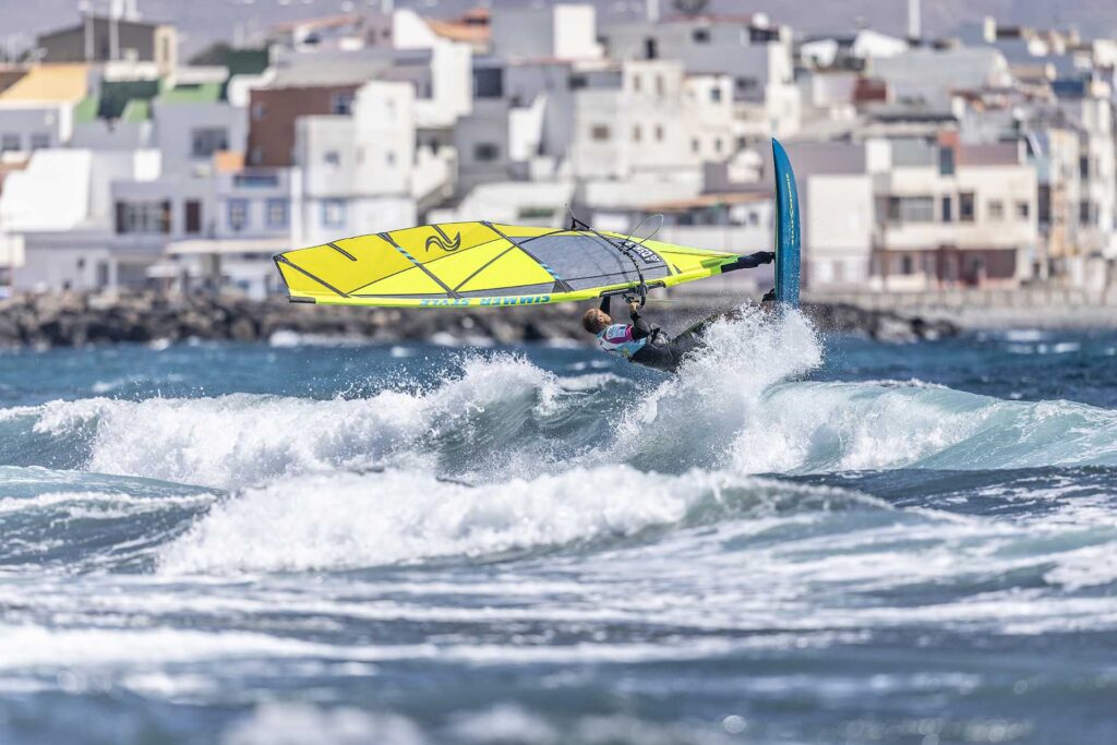 Windsurfer riding a bright yellow sail on breaking waves near a coastal town.