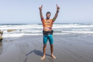 Man on a wet sandy beach, wearing an orange surfing jersey, arms raised in celebration with waves behind him.