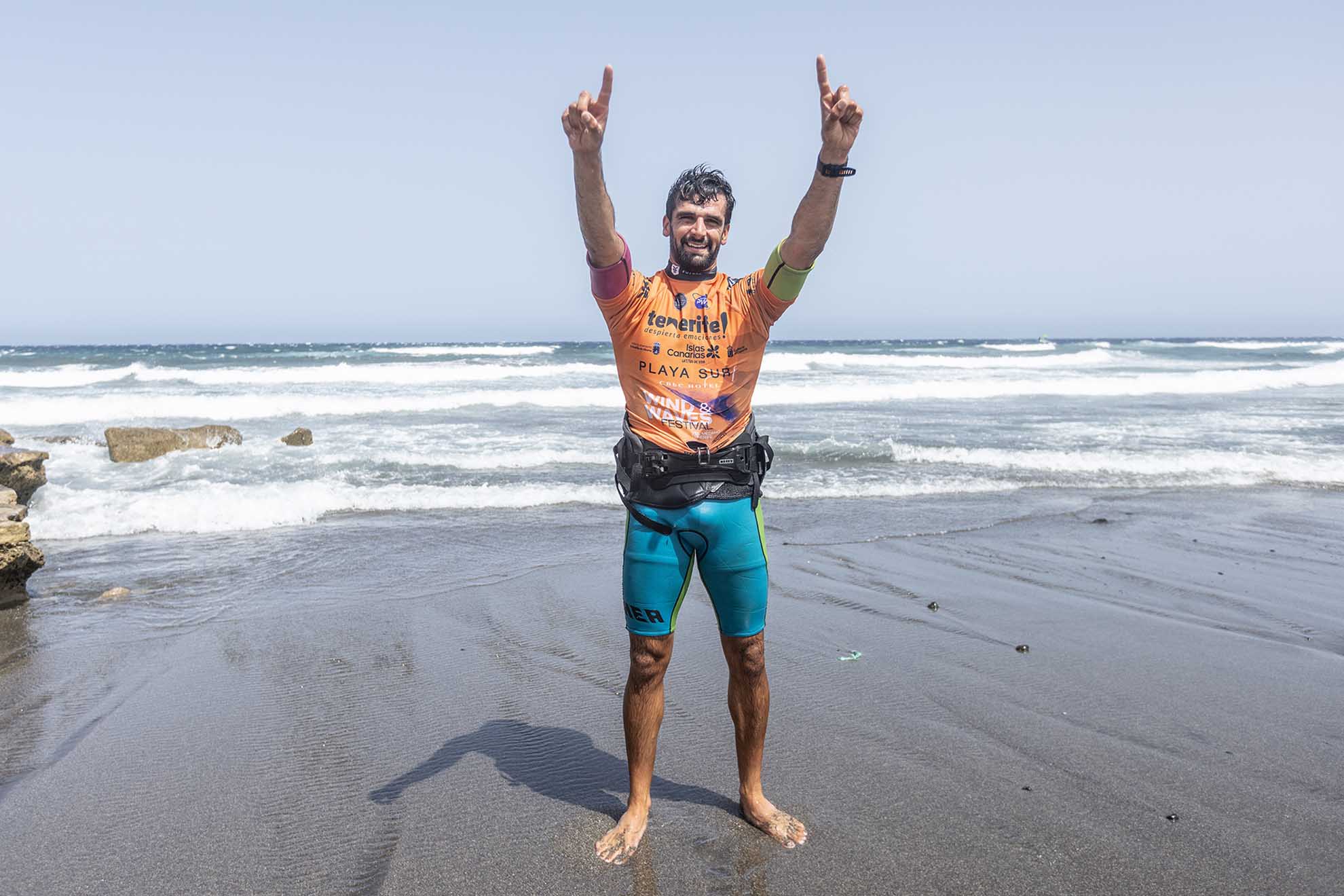 Man on a wet sandy beach, wearing an orange surfing jersey, arms raised in celebration with waves behind him.